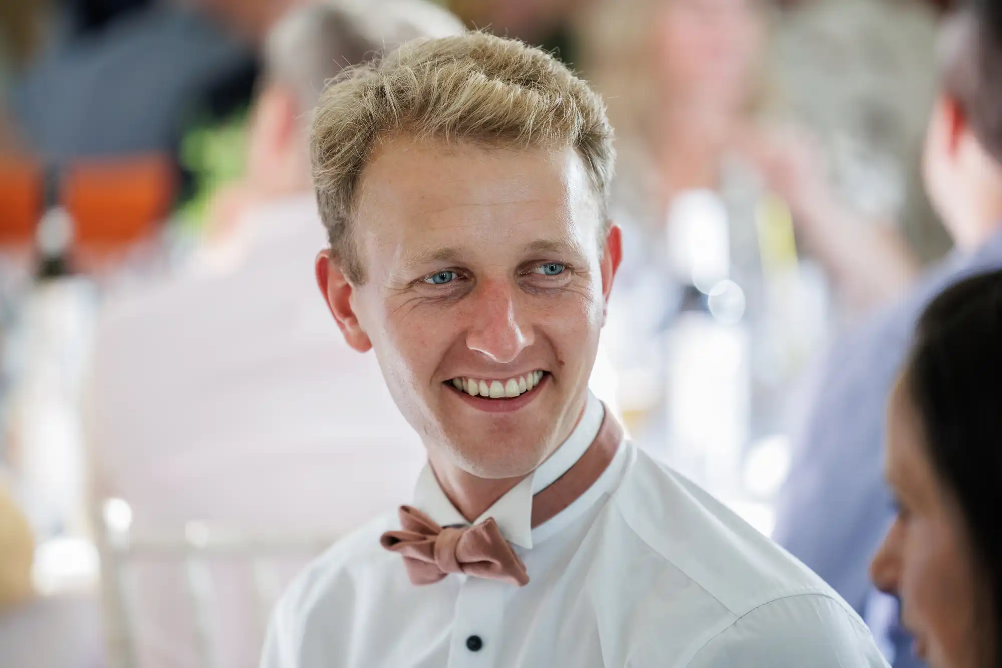 A young man with blond hair smiles whilst wearing a white shirt and a loosened tan bow tie around his neck. The background is softly blurred, showing other people and a bright, indoor setting, suggesting a celebratory or social event.
