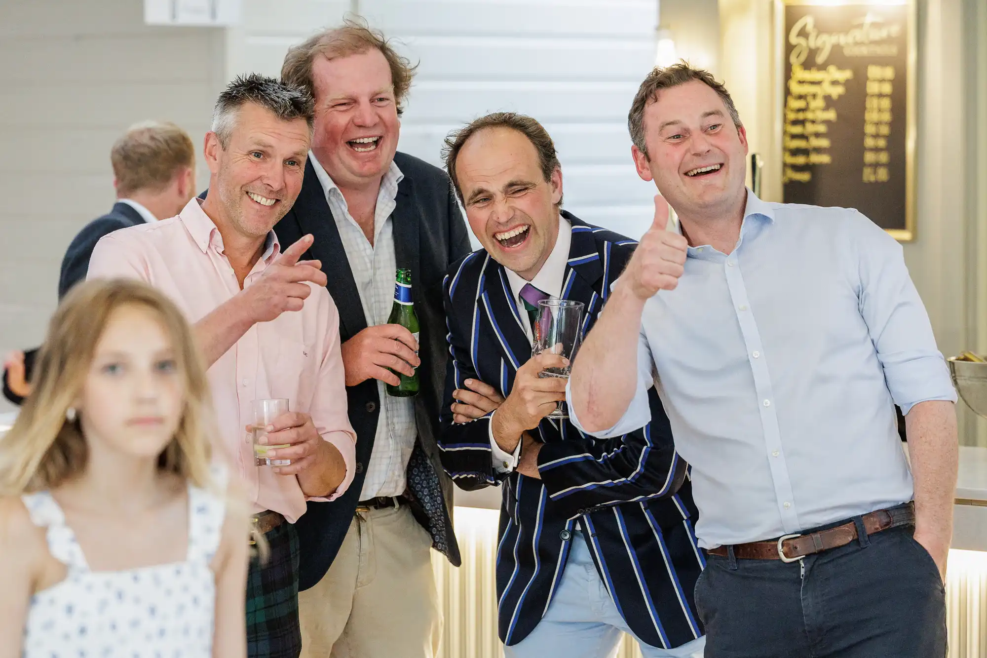 Four men dressed smartly, laughing and posing with drinks in hand; one wears a striped blazer, another holds a pint. They gesture enthusiastically at the camera. A young girl in a white dress stands in the foreground, slightly out of focus. Indoor setting, bright lighting.
