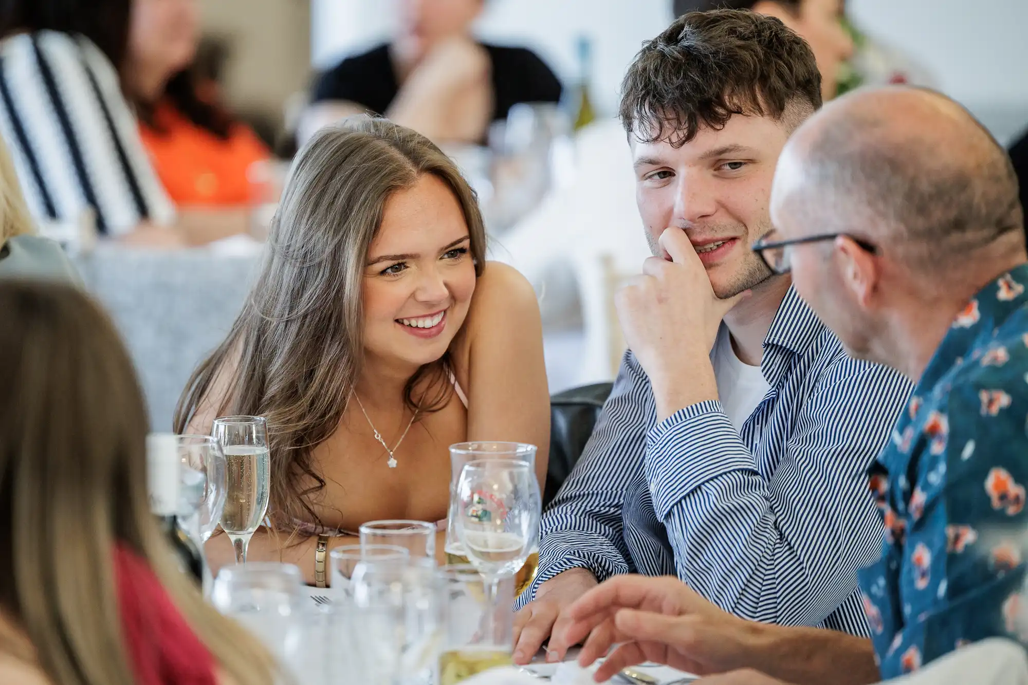 A young woman smiles and leans in to talk with a young man and an older man at a table set with wine glasses and plates. Others are blurred in the background, suggesting a lively social or celebratory event.
