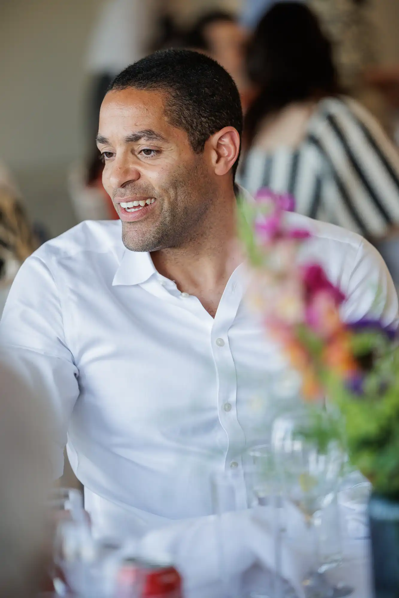 A man wearing a white button-up shirt smiles whilst sitting at a table. Colourful flowers and glassware are in the foreground, slightly out of focus. Other people and a softly lit background create a warm, social atmosphere.
