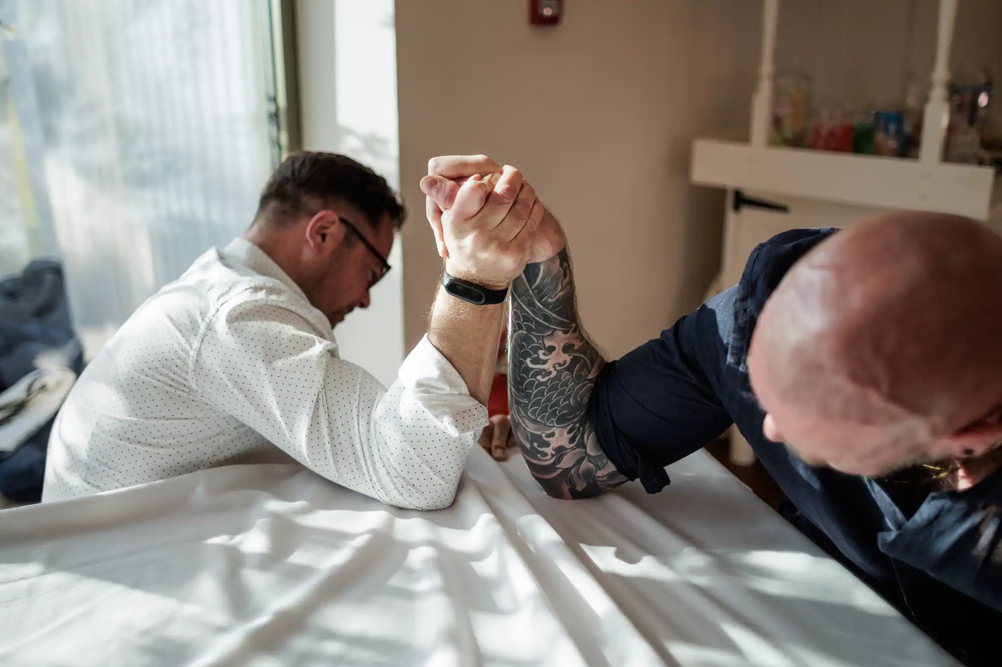 Two men arm wrestling on a table in a sunlit room. The man on the left wears glasses and a white shirt, while the man on the right has a bald head, a dark shirt, and a tattooed arm. Both are leaning in with effort, gripping each other’s hand tightly.