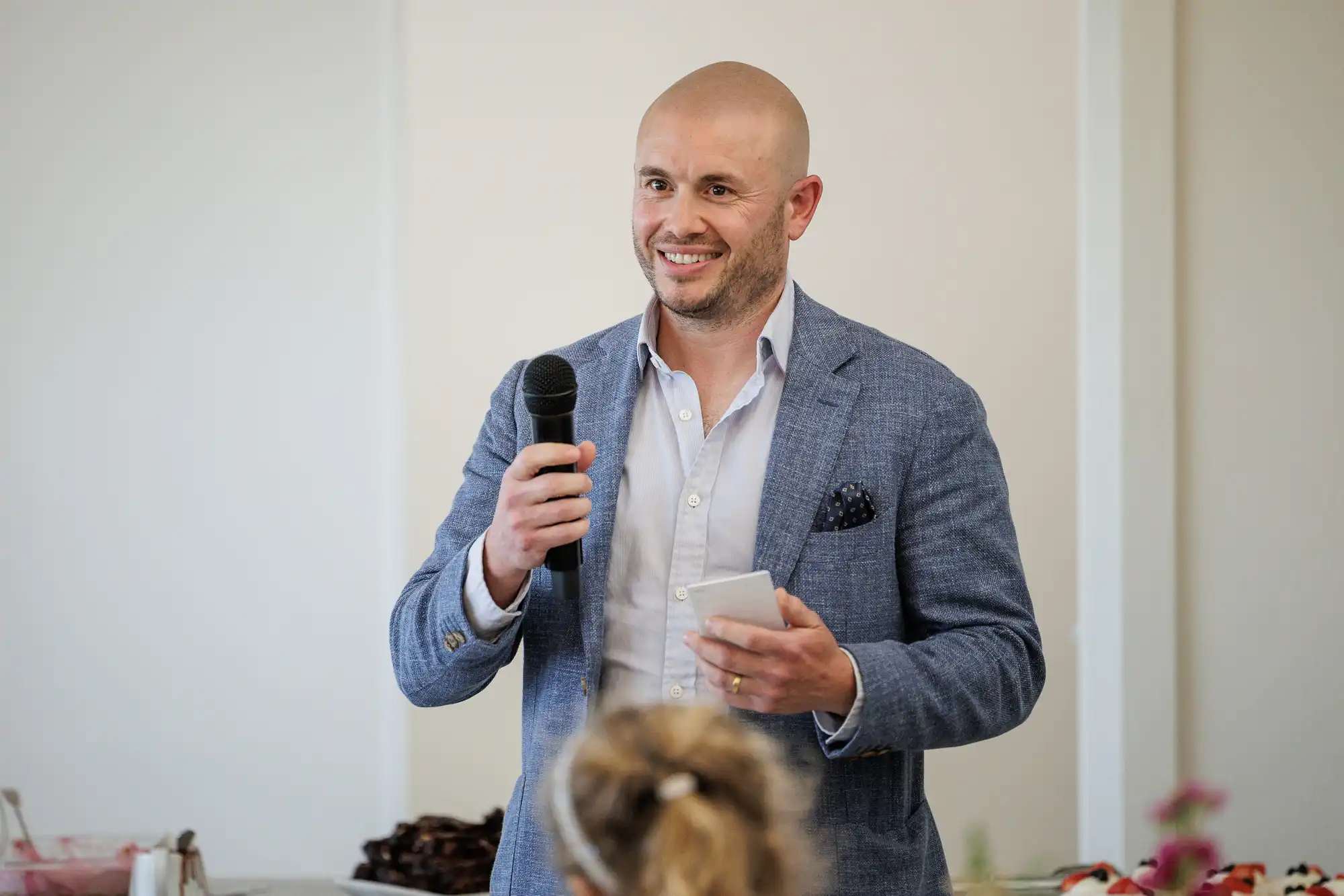 A smiling bald man in a blue blazer and white shirt holds a microphone and a notepad, speaking to an audience indoors. The background is softly lit and out of focus, with food visible on a table to the left.