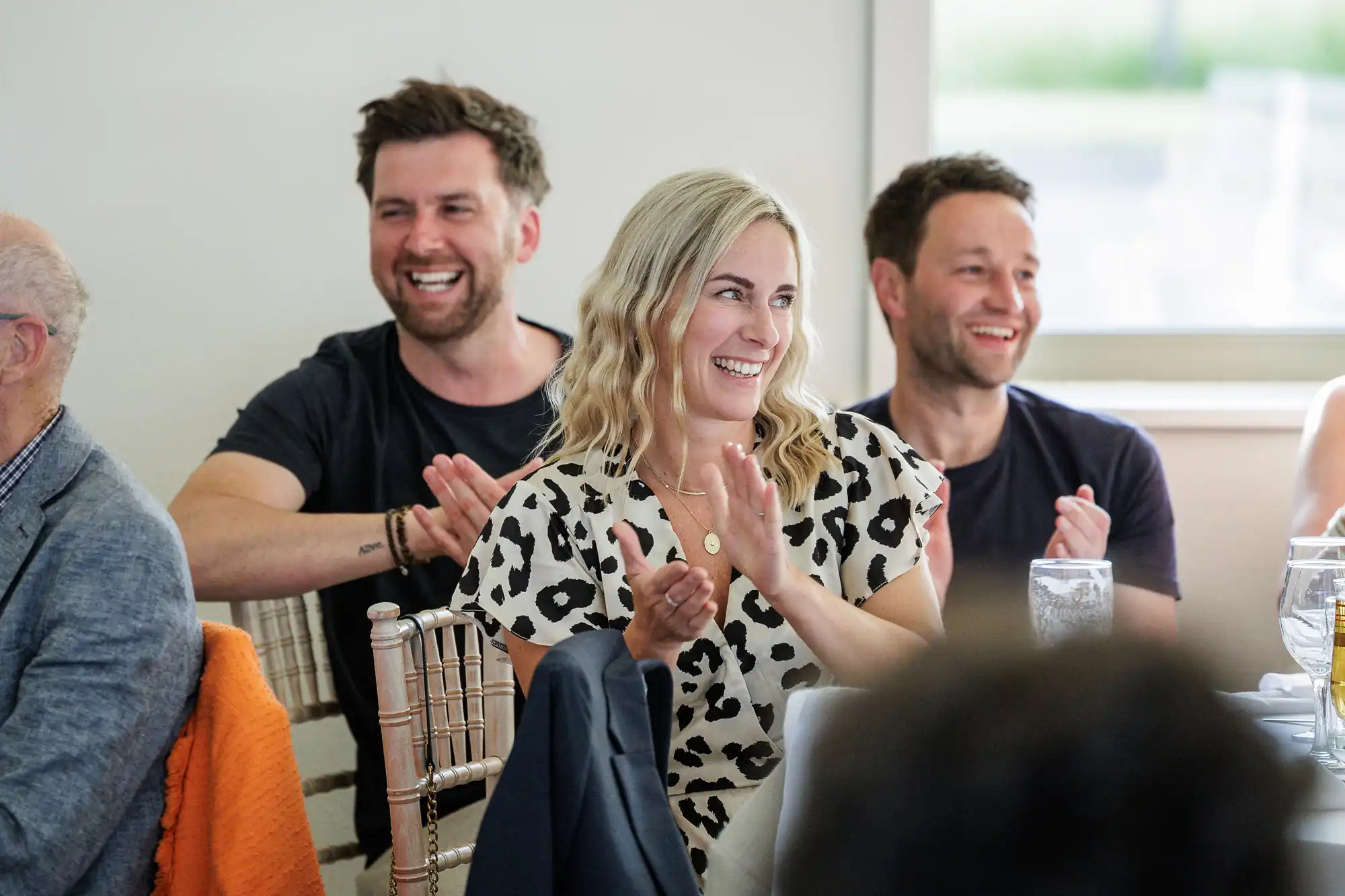 Three people sit together at a table, clapping and smiling during an indoor event. The woman in the centre has blonde, wavy hair and wears a white and black patterned top. Two men sit on either side of her, both with short hair and dark shirts.
