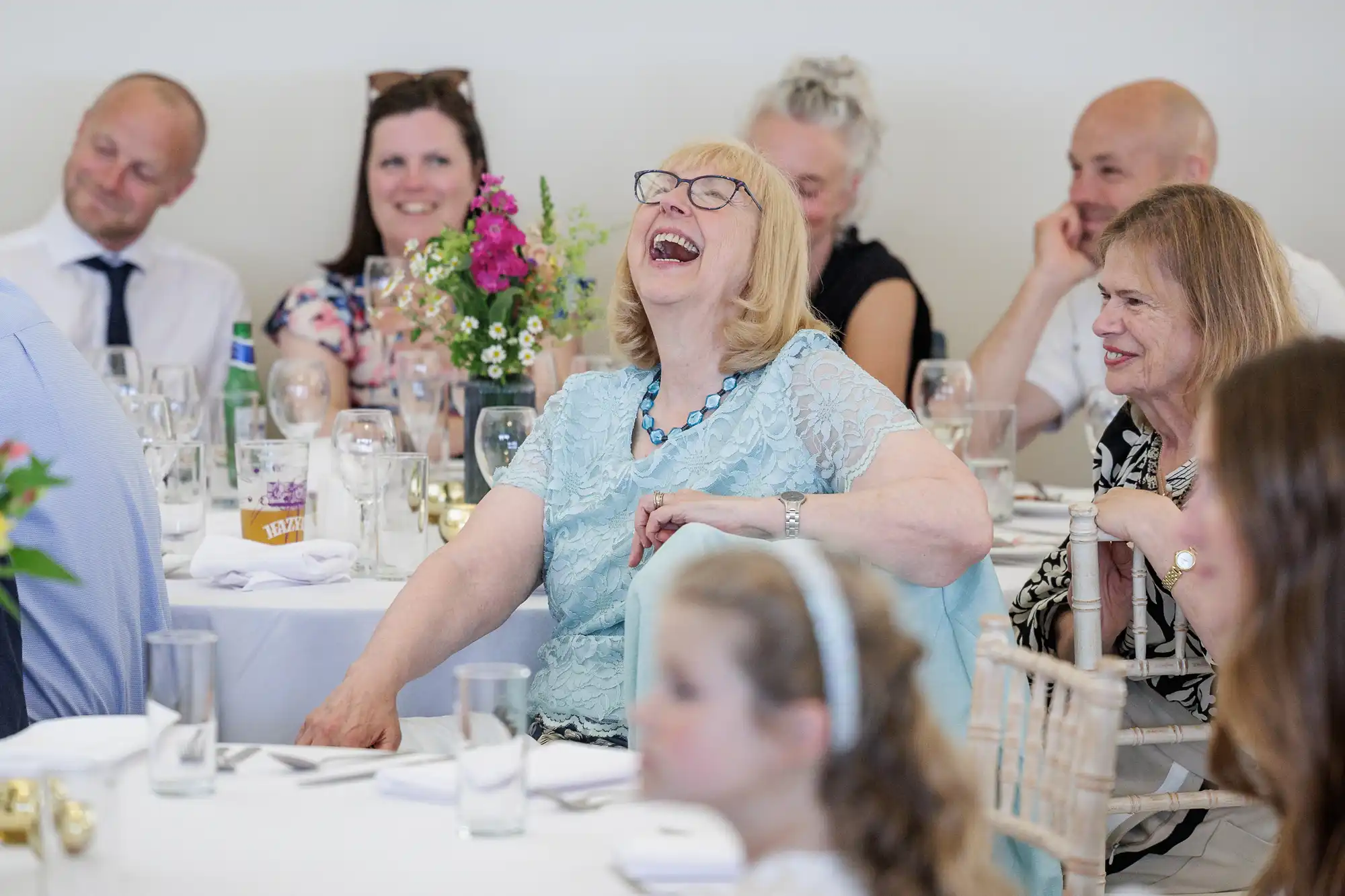 A joyful elderly woman in a light blue dress laughs heartily at a table set for a formal gathering, surrounded by smiling guests. Flowers and glassware decorate the tables, and the atmosphere is lively and cheerful.