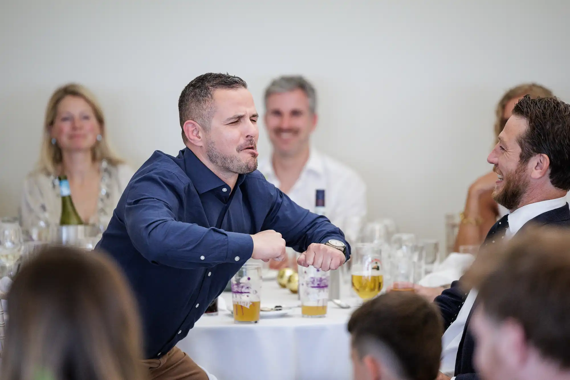 A man in a blue shirt stands by a table, making a playful pose with bent arms and clenched fists. He appears to be entertaining or joking, while others seated around him, including a man in a suit, laugh and smile. The setting looks like a social gathering or event.