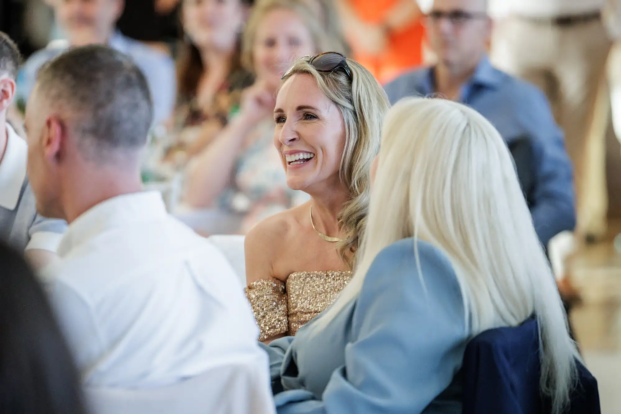A blonde woman in a sparkly off-the-shoulder dress sits smiling among a crowd at an indoor event. She wears sunglasses on her head. People around her, some blurred, appear to be engaged and listening attentively.