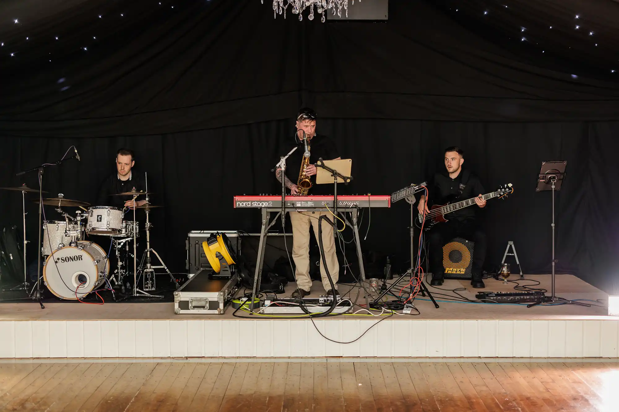 Three musicians perform on stage under a black, starry canopy. The drummer is on the left, a keyboard player with a microphone stands centre, and the bassist sits right. Various instruments, cables, and equipment are visible around them on the wooden stage.