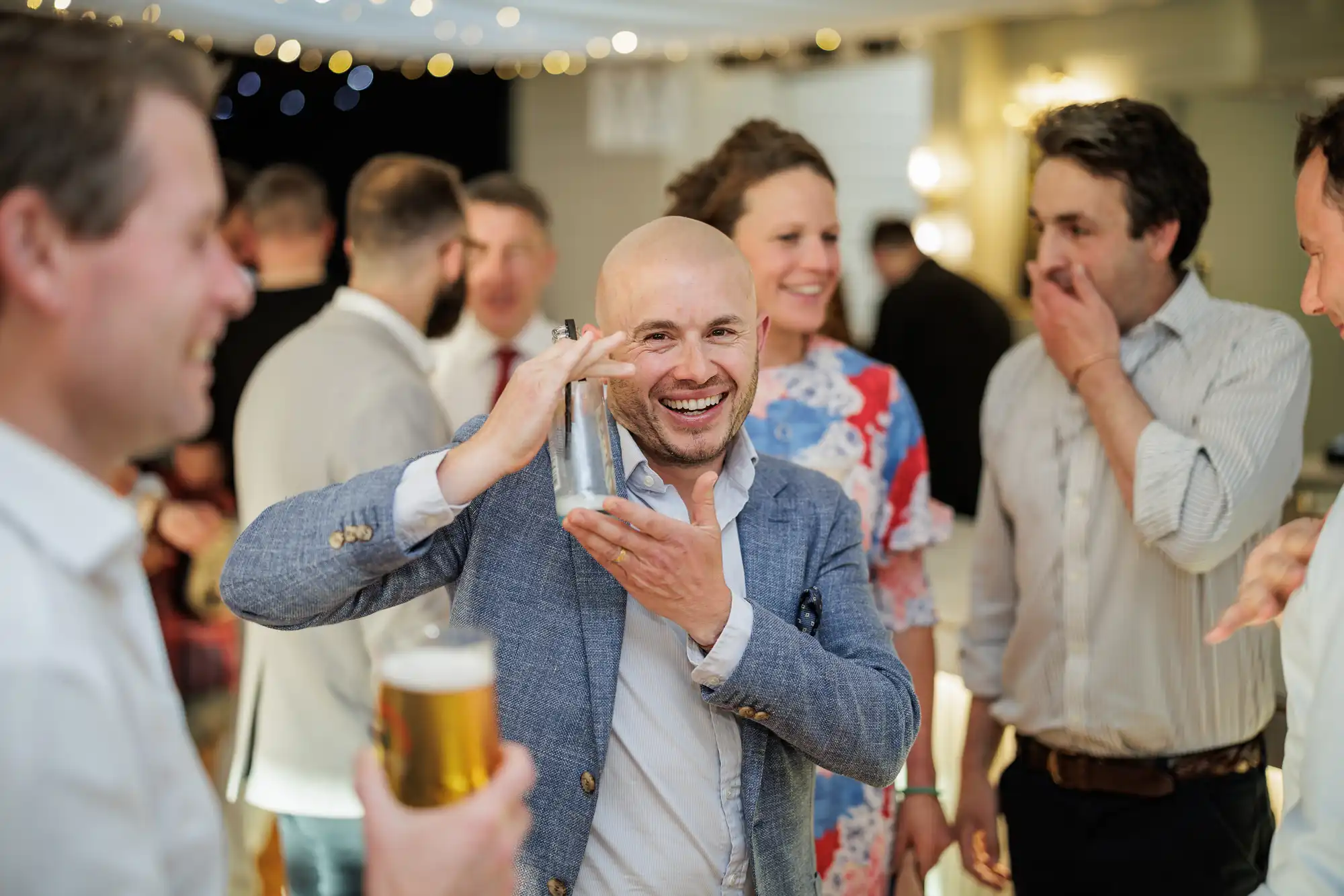 A smiling bald man in a blue blazer shakes a cocktail shaker at a lively indoor gathering. Around him, well-dressed people laugh and chat. Warm lights and fairy lights create a festive atmosphere. One man holds a pint of beer in the foreground.