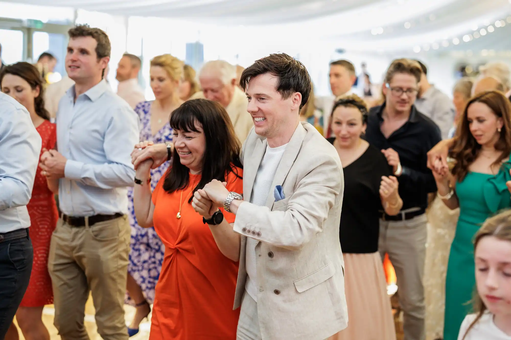 A group of people in colourful, semi-formal attire smile and hold hands while dancing closely together indoors under fairy lights, suggesting a lively celebration or wedding reception. The atmosphere is joyful and energetic.