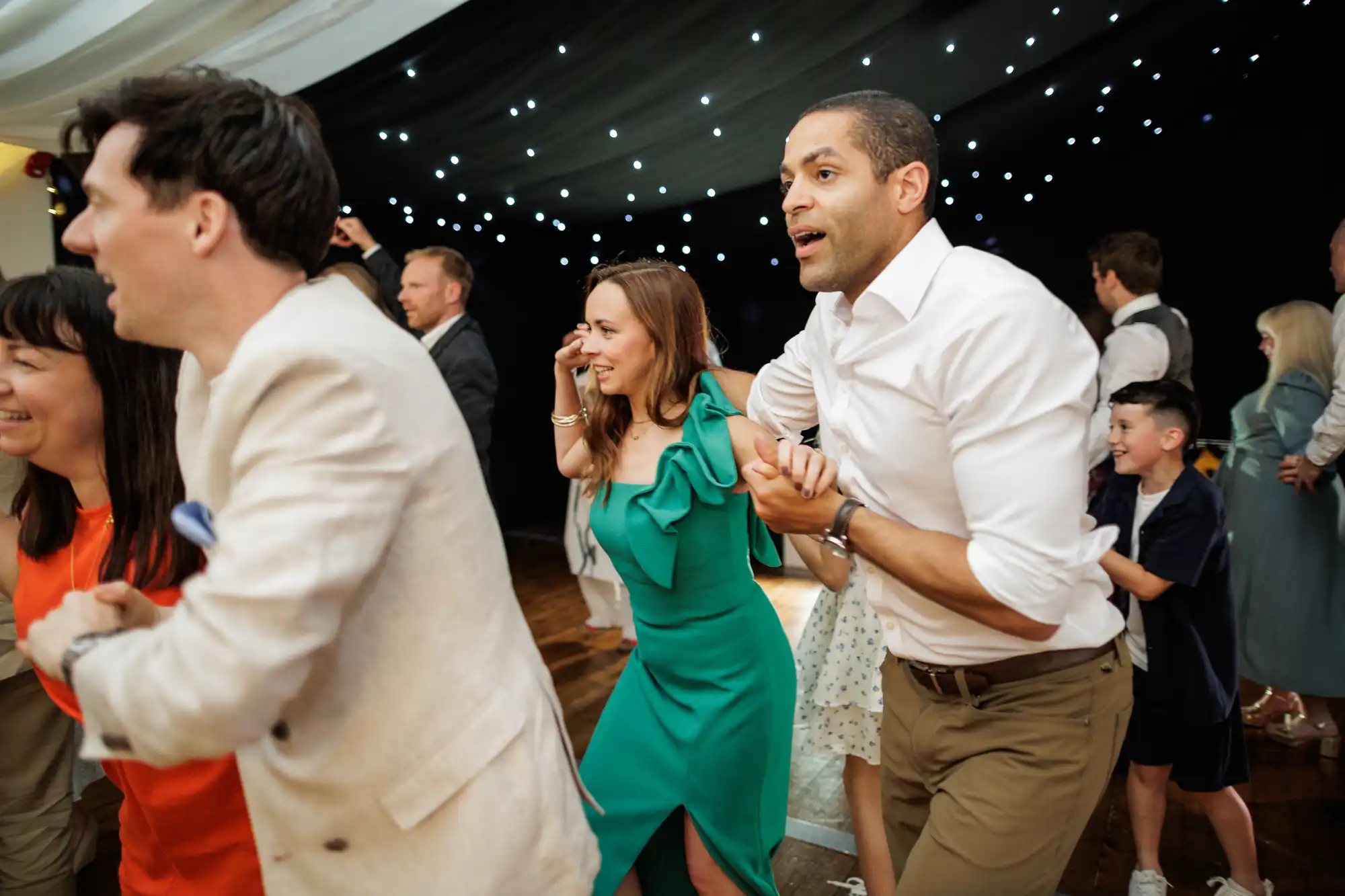 A group of people, dressed in semi-formal attire, are laughing and holding hands whilst dancing in a conga line at an indoor event with fairy lights and a starry black canopy above them. Everyone appears joyful and engaged in the celebration.