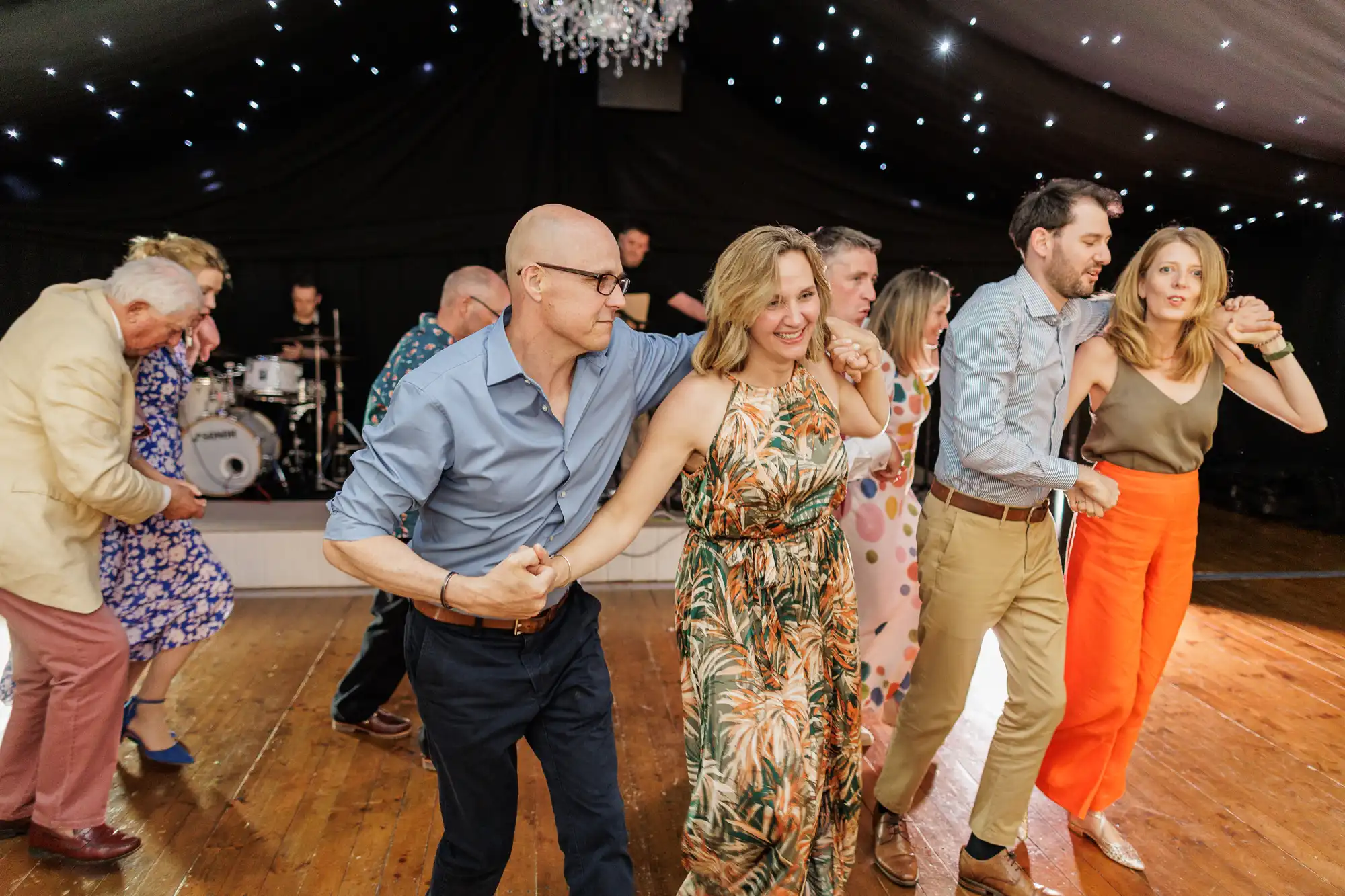 A group of adults hold hands and dance energetically in a line on a wooden floor under a dark, starry-lit ceiling. Everyone is smiling and dressed in colourful, festive outfits. A live band with drums is visible in the background.
