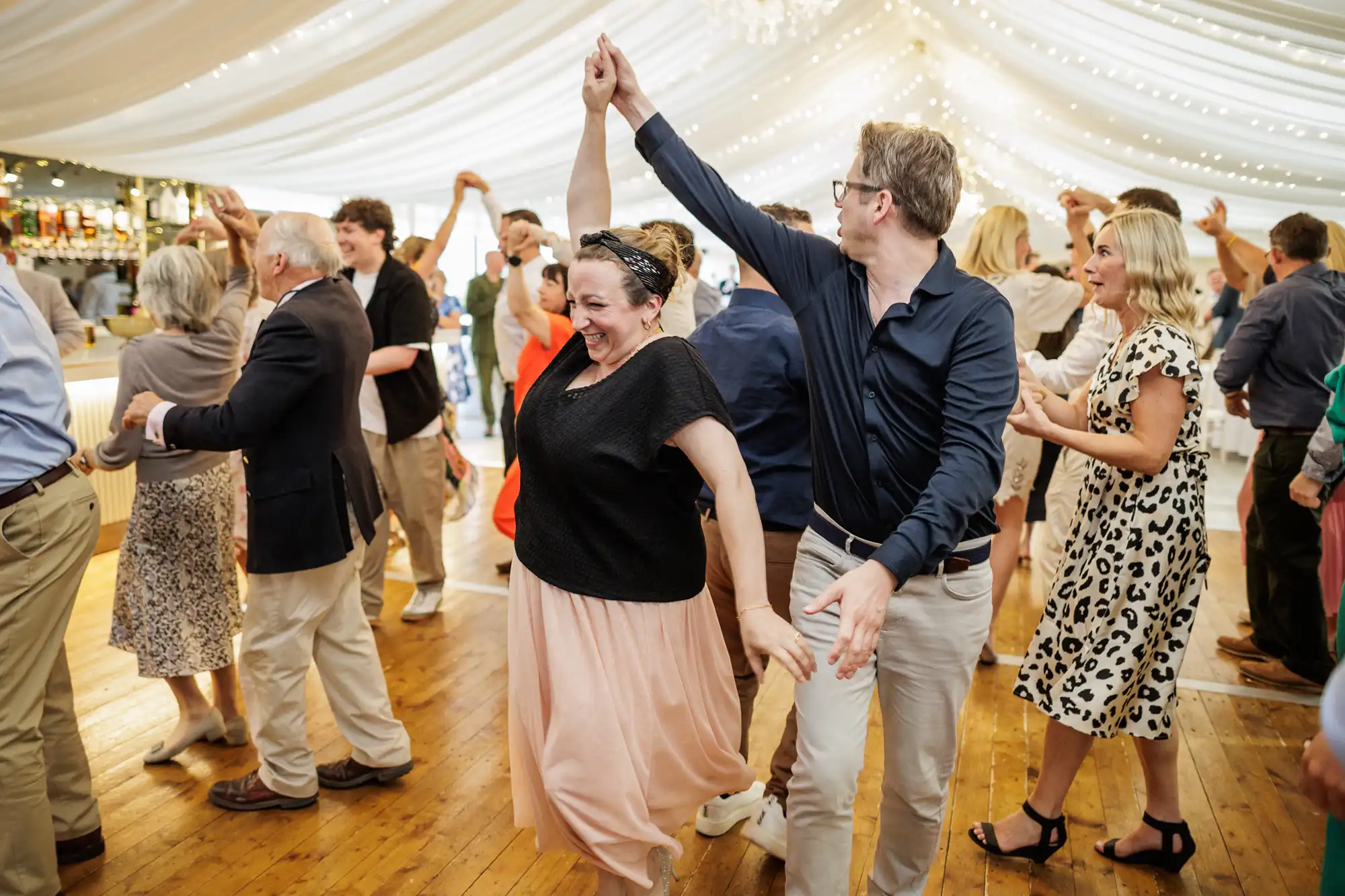 A joyful group of people dance together under a white marquee with fairy lights. A smiling woman in a black top and peach skirt twirls with a man in a navy shirt and light trousers. Others around them laugh, clap, and enjoy the lively atmosphere on the wooden floor.