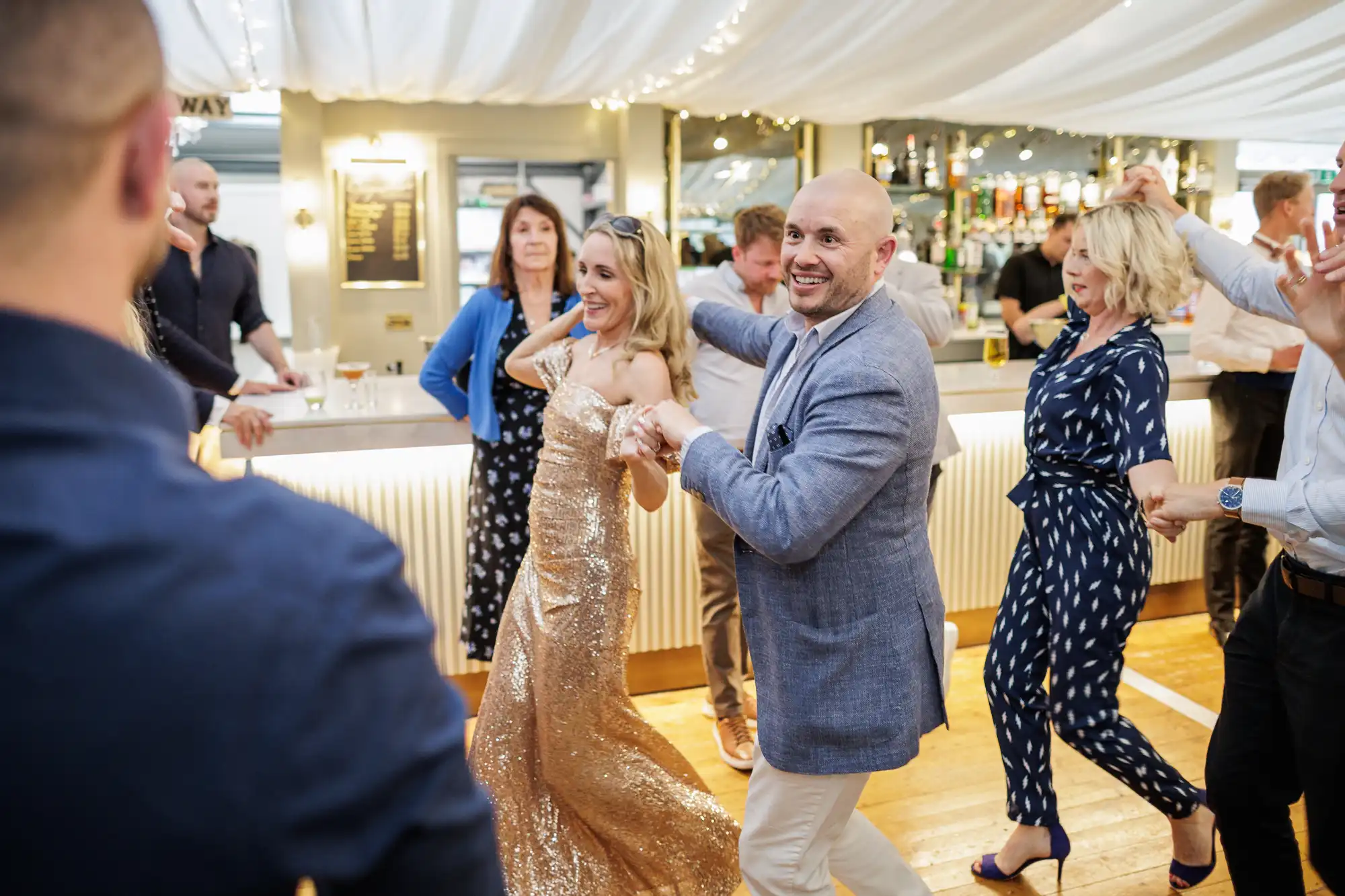A joyful group of people dance indoors; a woman in a sparkling gold dress and a smiling bald man in a light suit lead the queue. Others, dressed smartly, hold hands and follow. The setting is bright, with a bar in the background and fairy lights overhead.