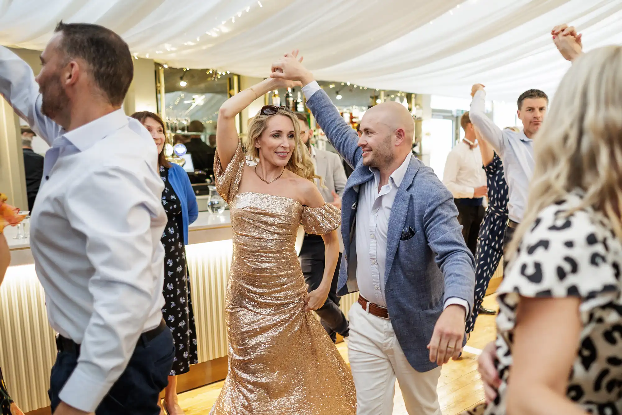 A man in a blue blazer and white trousers spins a woman in a glittery gold dress as they dance at a festive indoor event, surrounded by other couples dancing under white drapes and fairy lights.