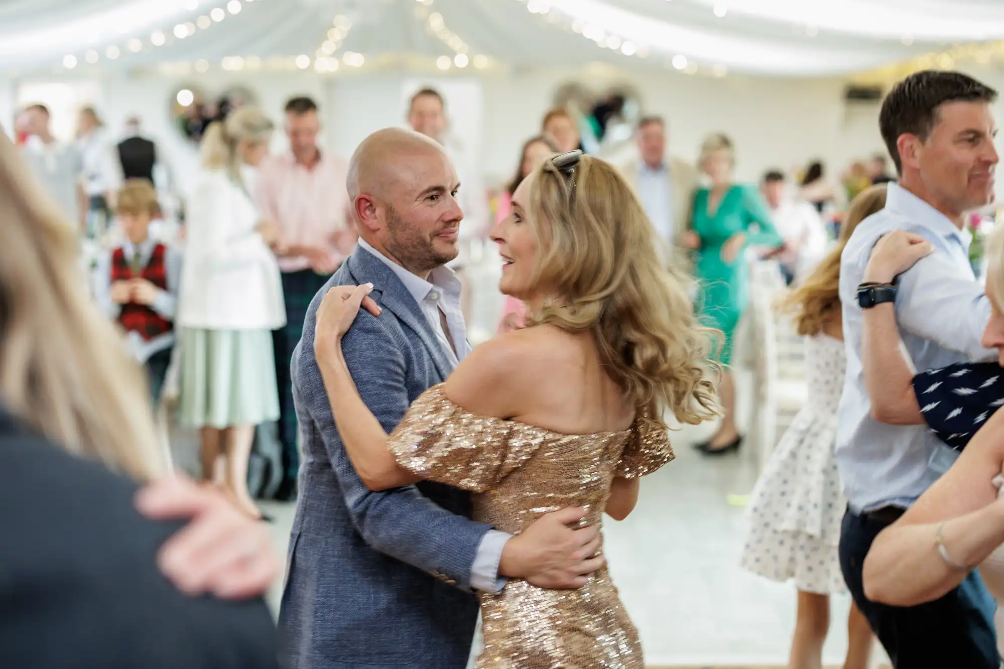 A man in a light blue suit jacket dances with a woman in a sparkling gold off-the-shoulder dress at a lively indoor event. Other couples dance around them under a ceiling strung with white lights. The background is blurred with guests talking and mingling.