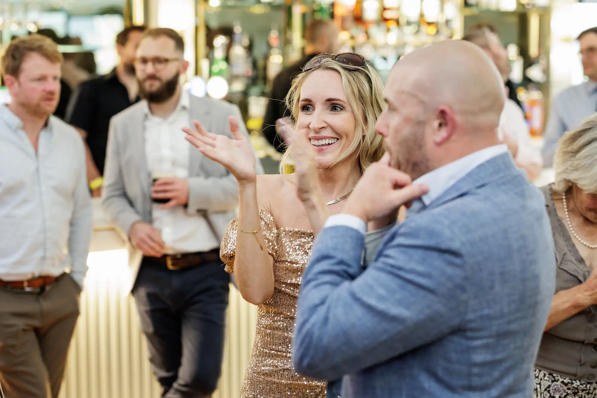 A smiling woman in a sparkly gold dress claps her hands at a lively indoor event. She is surrounded by casually dressed people, some talking and holding drinks, with a well-lit bar in the background. The mood is joyful and social.