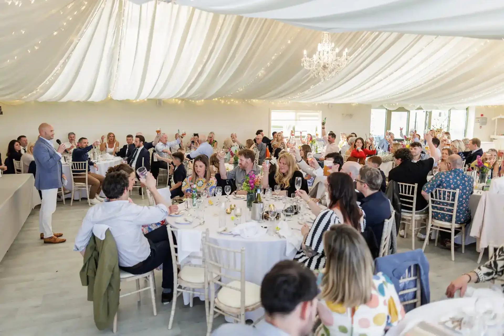 A wedding reception in a bright, elegant marquee at Runningburn wedding venue, with white draped ceiling and chandeliers. Guests sit at round tables, toasting with champagne as a person in a blue blazer gives a speech at the front. The atmosphere is festive and joyful.