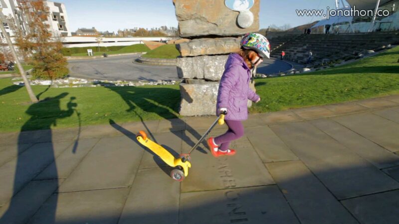 A child in a purple jacket and helmet pulls a yellow scooter along a paved walkway on a sunny day. A stone structure and grassy area are in the background - Videographer in Edinburgh testing a Steadicam gimbal with 3 year-old Anabelle scooting around Holyrood!
