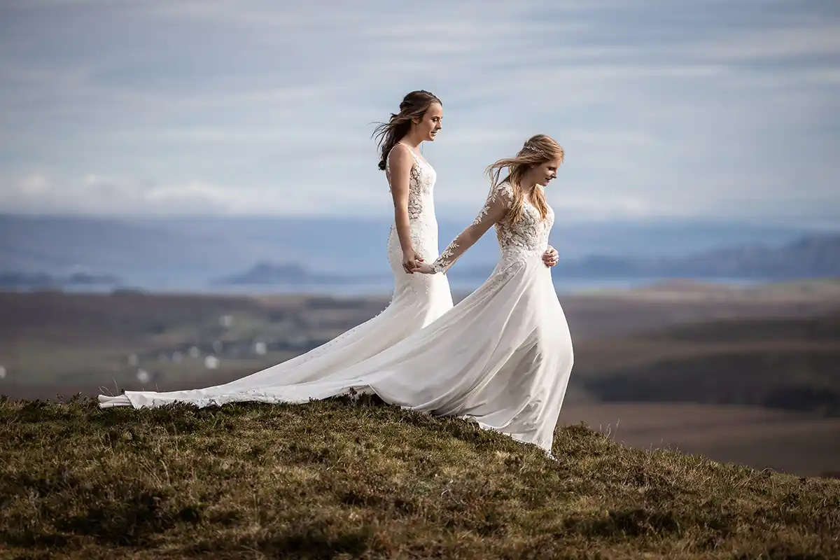 Two brides in long, elegant white wedding dresses hold hands whilst walking on a grassy hilltop. The flowing dresses trail behind them. The landscape features distant hills, water, and a cloudy sky, creating a serene and romantic atmosphere. Scottish elopement photographer at The Quiraing, Isle of Skye.