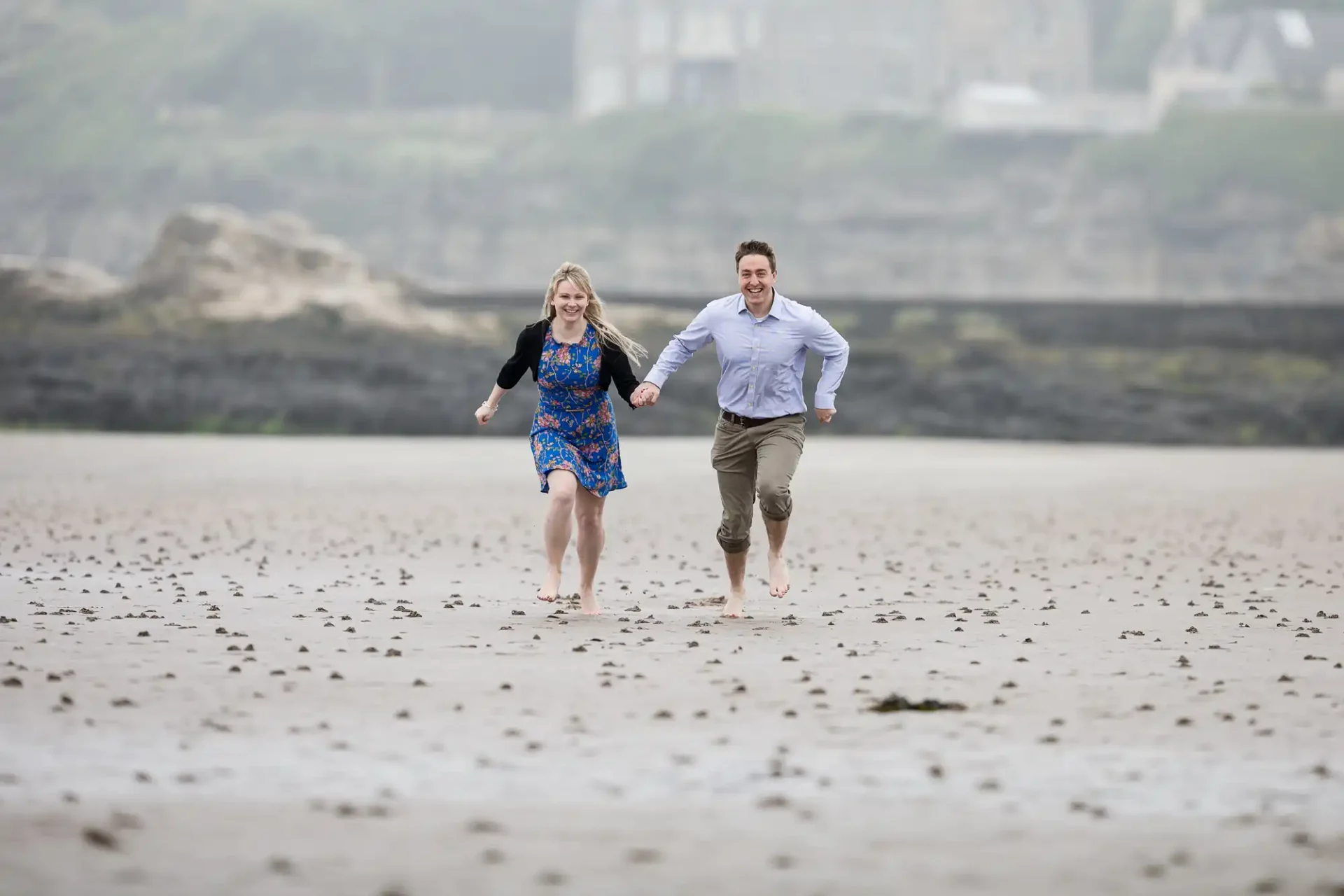 A man and a woman run barefoot on a sandy St Andrews Beach, holding hands and smiling. The woman wears a blue floral dress and black cardigan; the man wears rolled-up khaki trousers and a light blue shirt. Rocky cliffs and blurred buildings appear in the background.