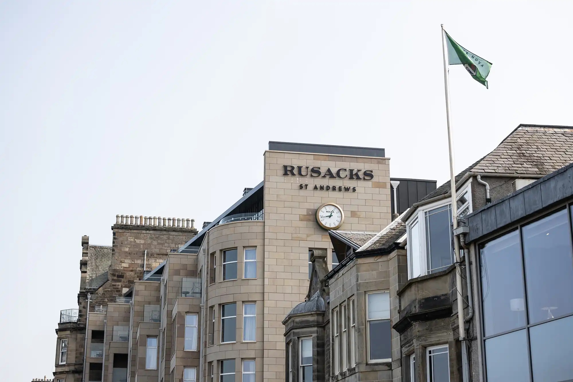 A beige stone building with a sign reading "Rusacks St Andrews" and a round clock on its facade. The structure has classic and modern architectural features, and a green and white flag flies on a pole to the right. The sky is clear and pale.