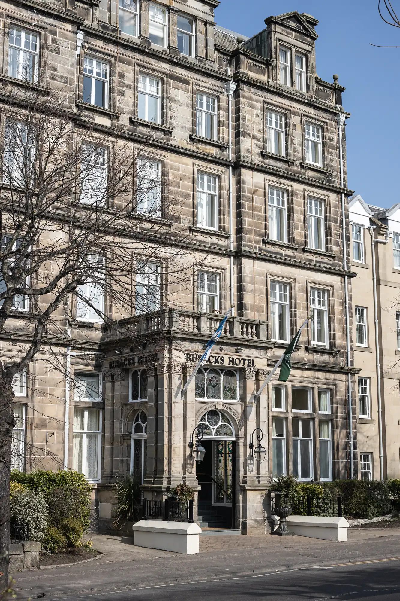 A grand stone building with arched windows and an ornate entrance, featuring a sign reading “Rudding’s Hotel.” Two flags hang above the door, and bare trees line the pavement in front. The sky is clear and blue, suggesting a calm day.