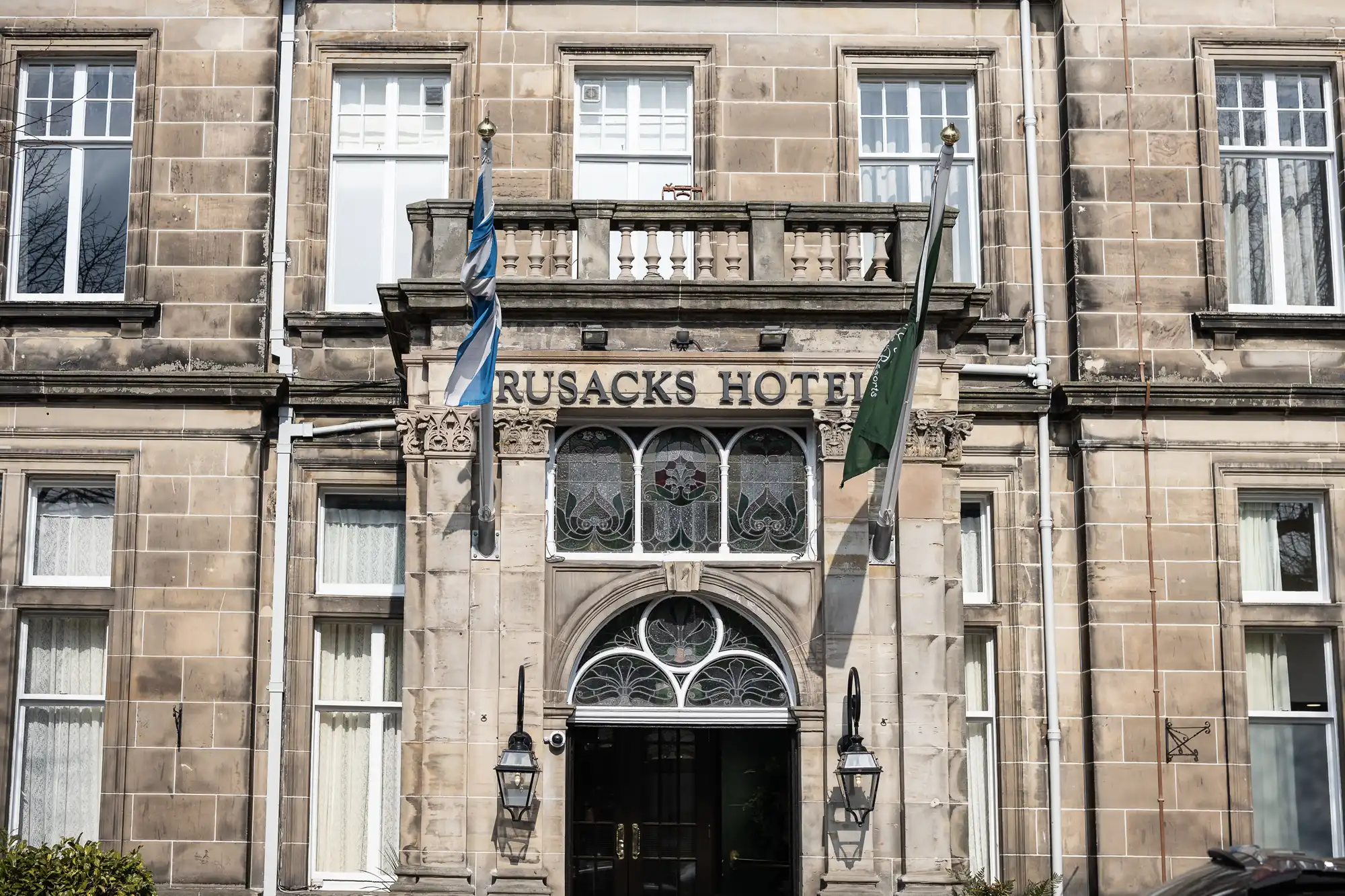The image shows the stone façade of Rusacks Hotel. There is an arched entrance with ornate glass windows above the doorway, flanked by two lamps. Two flags hang outside the entrance, and large windows are visible on both sides of the building.
