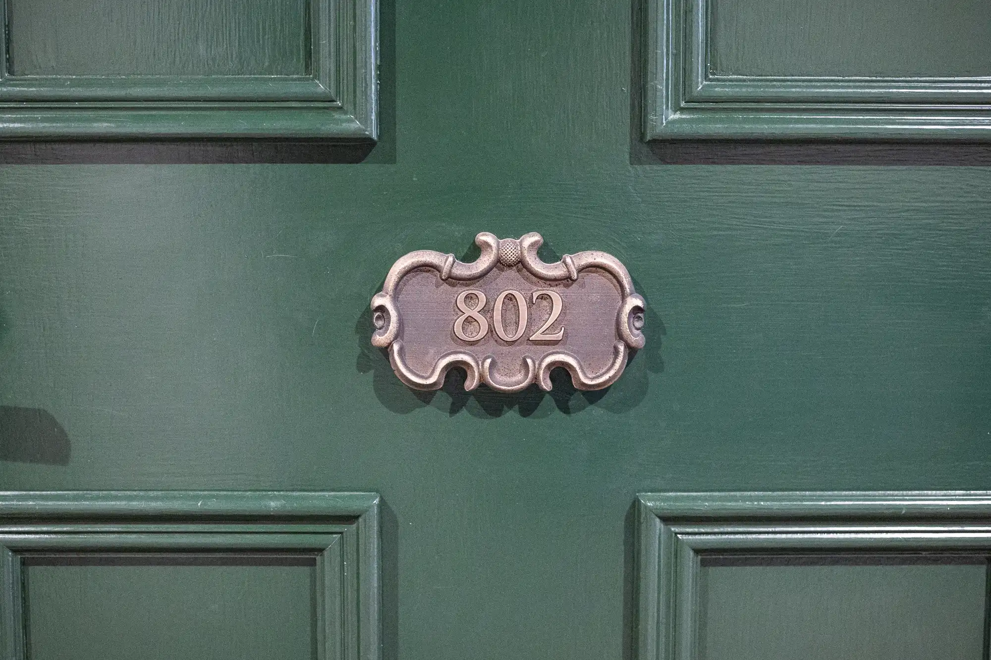 A close-up of a dark green door featuring a decorative, ornate metallic plaque with the number "802" engraved in gold at the centre. The door has rectangular panel detailing surrounding the plaque.