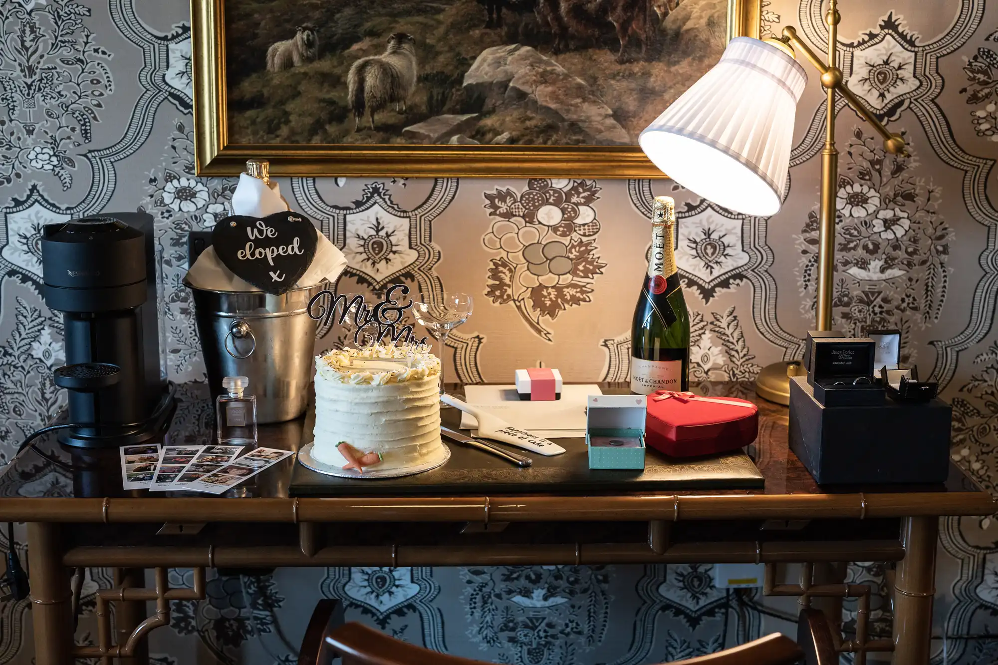 A decorated desk displays a white iced cake with “Mr & Mrs” toppers, champagne in an ice bucket, wine glasses, a lamp, Polaroid photos, small gift boxes, and a red heart-shaped box, set against vintage wallpaper and a framed sheep painting.