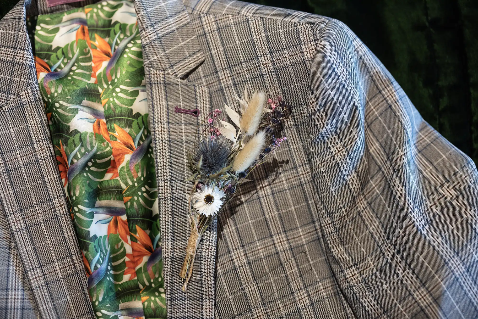 A grey checked suit jacket with a colourful tropical leaf and flower-patterned lining. The lapel has a rustic buttonhole made of dried flowers, grasses, and thistles attached with twine. The background is dark green.