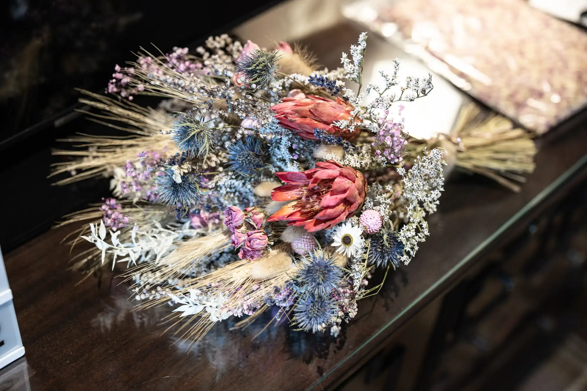 A bouquet of dried flowers, including pink protea, lavender sprigs, wheat stalks, and various small white and purple blossoms, lies on a dark wooden surface with blurred objects in the background.