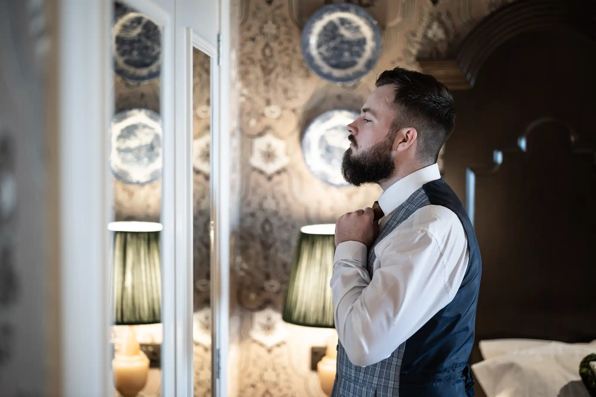 A man with a beard in a dress shirt and checked waistcoat adjusts his tie in front of a mirror in a stylishly decorated room with patterned wallpaper, decorative plates, and two lit lamps on either side of the mirror.
