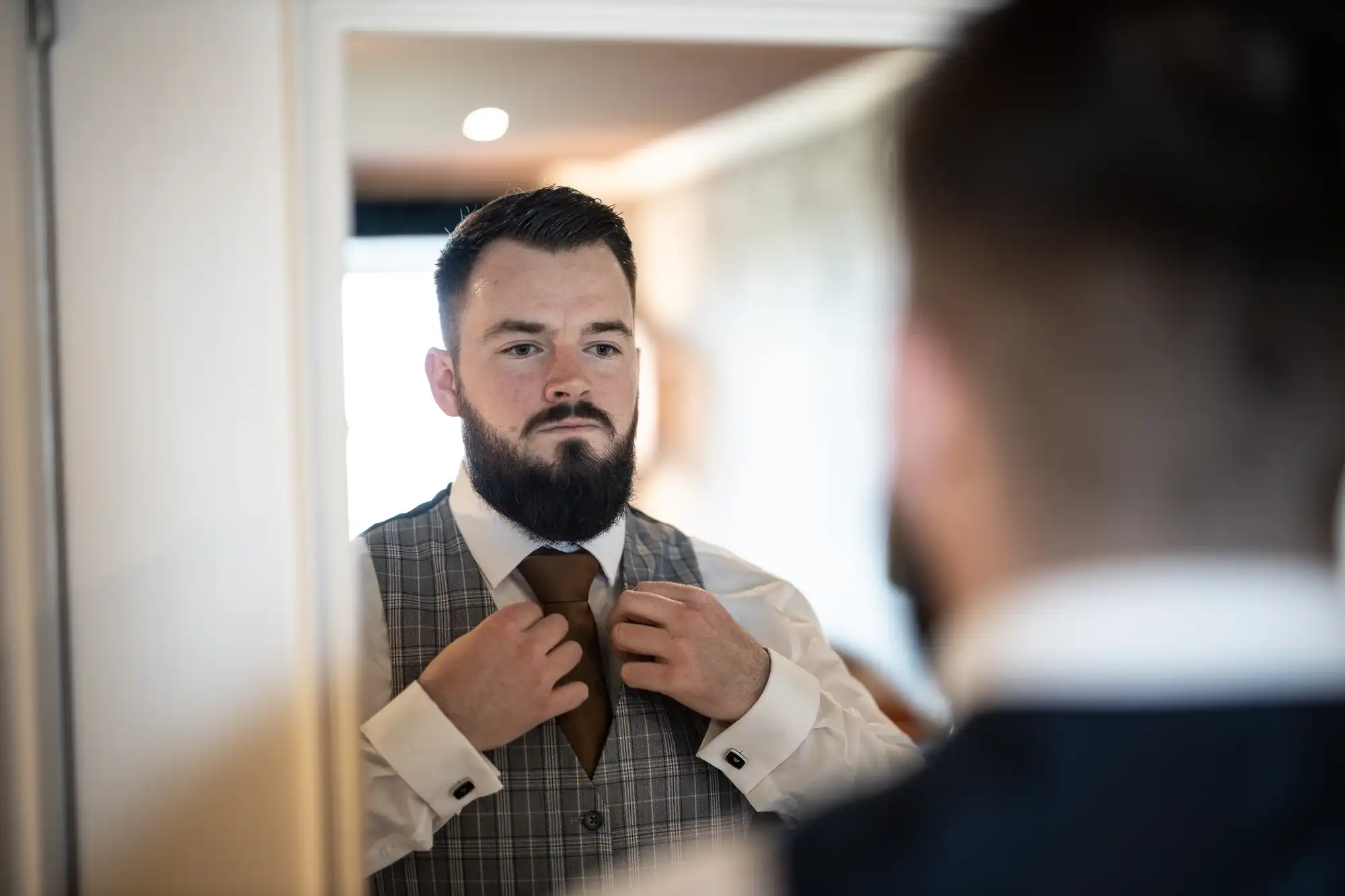A bearded man wearing a white shirt and checked waistcoat stands in front of a mirror, adjusting his brown tie. His reflection is visible as he focuses intently on straightening the tie in preparation, suggesting a formal or special occasion.