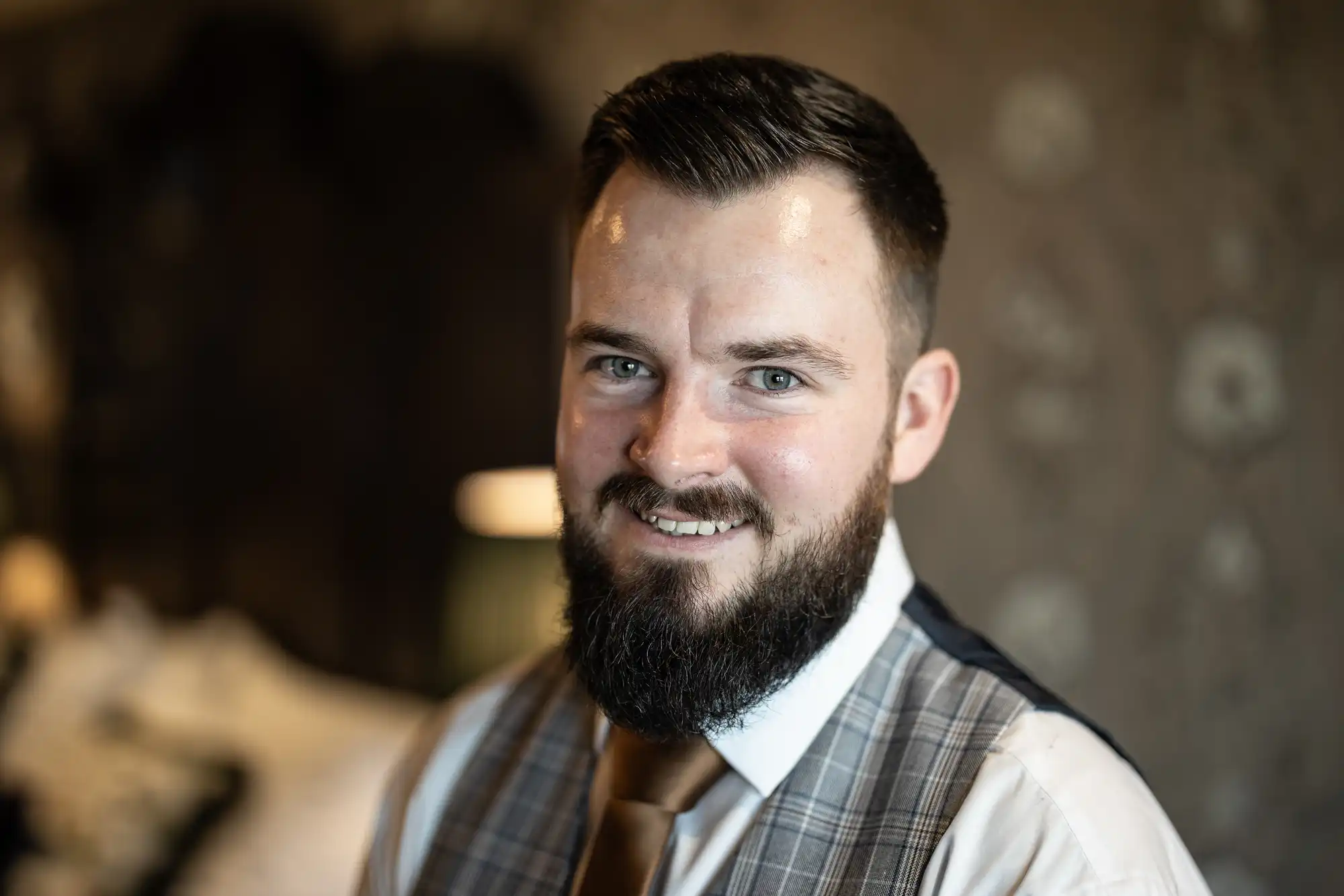 A man with short dark hair and a full beard smiles at the camera. He is wearing a white shirt, brown tie, and a grey checked waistcoat. The background is softly blurred with neutral tones, bringing focus to his face and upper body.