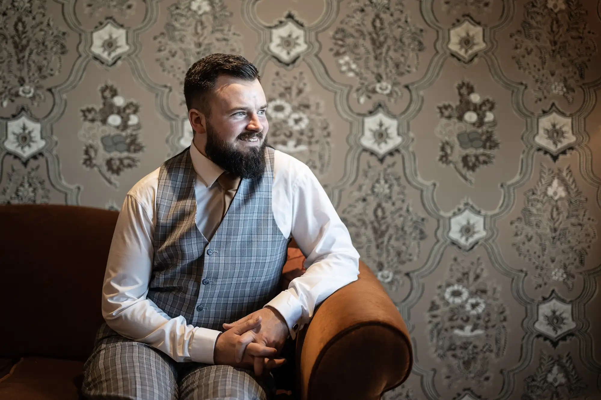 A man with a beard and slicked hair, wearing a grey checked waistcoat, white shirt, and brown tie, sits smiling on a brown sofa. The background features ornate, vintage-style floral wallpaper in muted tones. Light shines on him from the left.