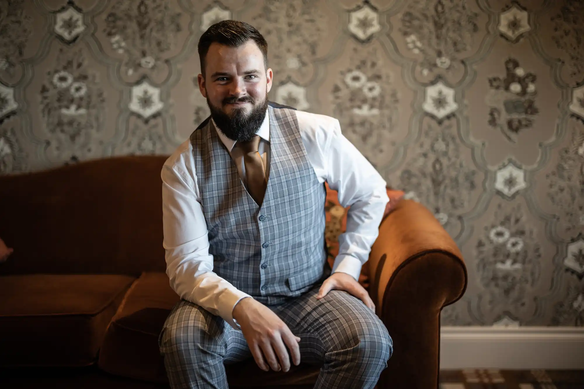 A bearded man with dark hair sits on a brown sofa. He wears a white shirt, brown tie, and a grey checked waistcoat and trousers. The background features ornate, patterned wallpaper in muted tones. He looks at the camera and smiles slightly.