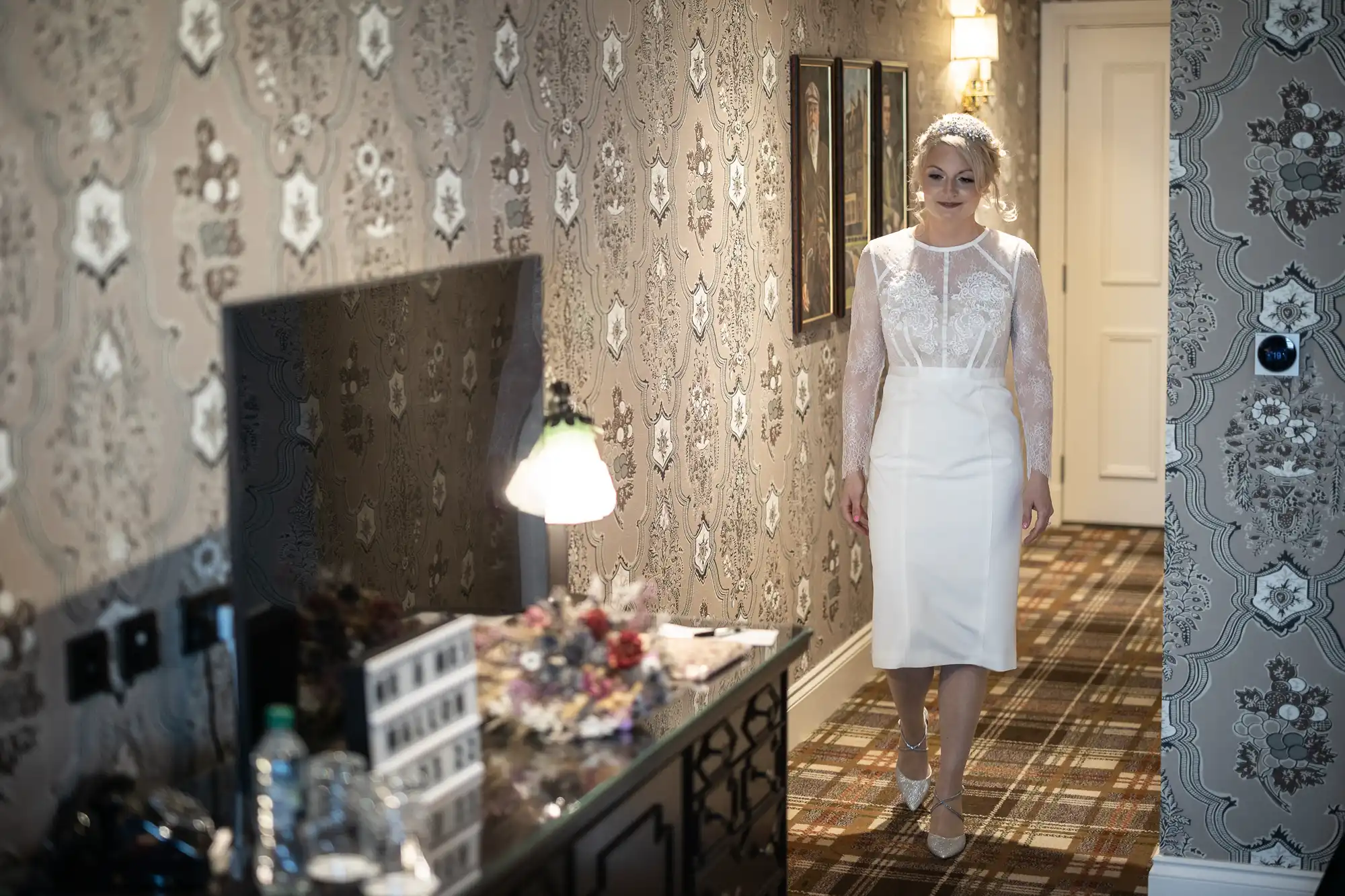 A woman in a white, knee-length bridal dress with lace sleeves walks down a patterned corridor with floral wallpaper and a checked carpet. She has a jewelled headpiece and is looking ahead. A table with a lamp, flowers, and framed items sits to her left.