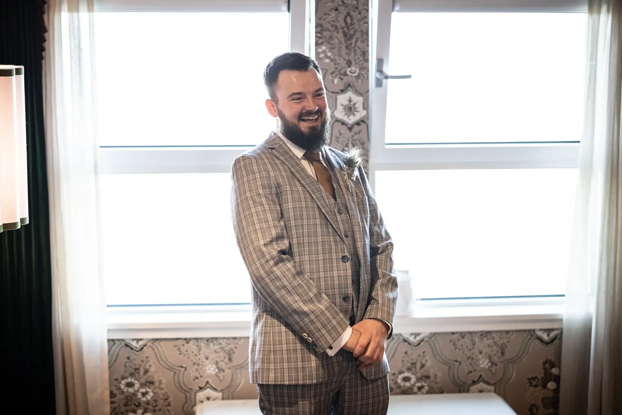 A bearded man wearing a grey checked suit with a tie smiles warmly, standing indoors in front of two large, bright windows with sheer curtains. Ornate patterned wallpaper is visible behind him, and soft natural light fills the room.