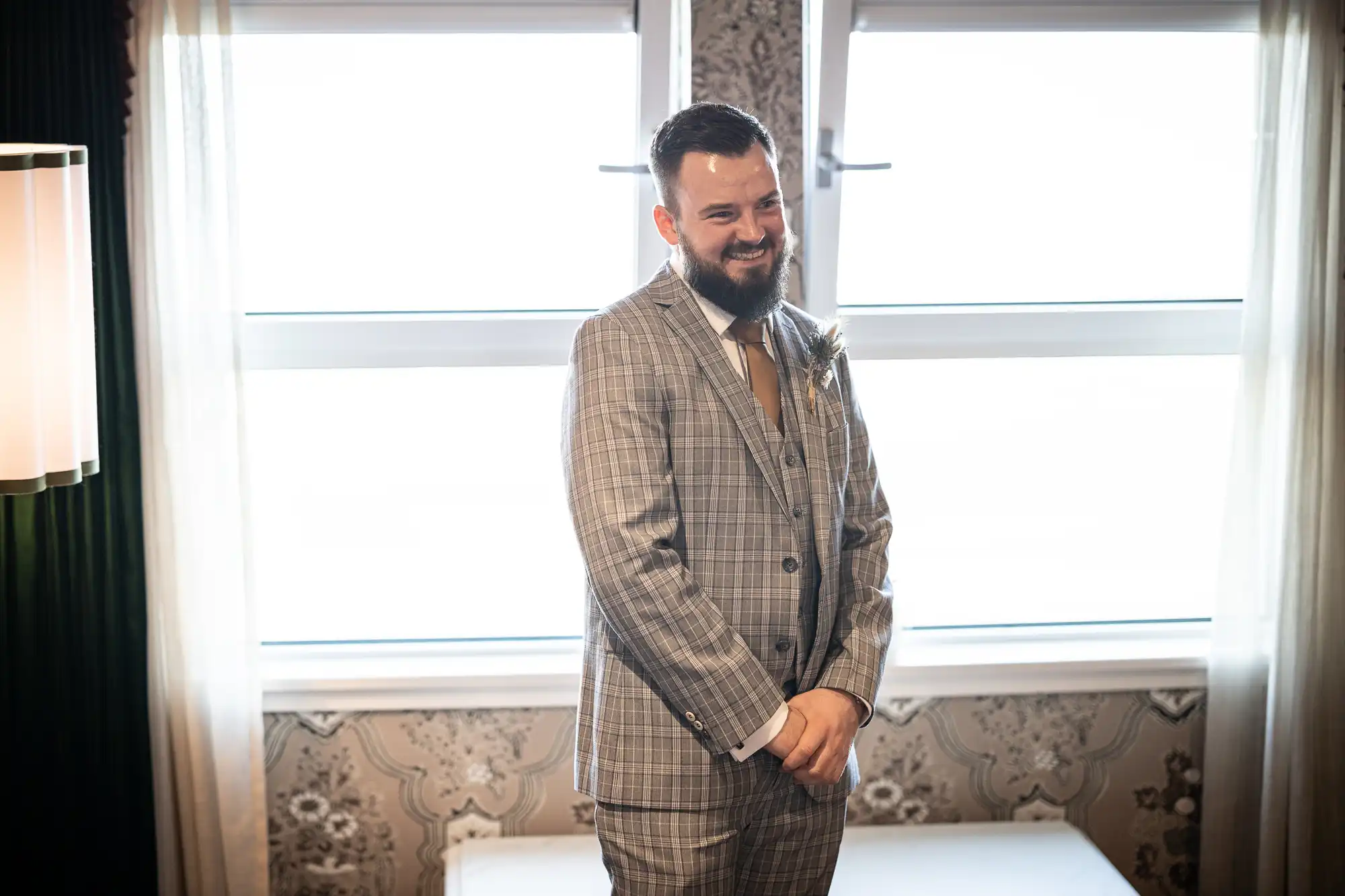 A man with a beard, wearing a light grey checked suit and brown tie, stands smiling in front of two bright windows with white curtains. The room has patterned wallpaper and a modern wall lamp is visible on the left.
