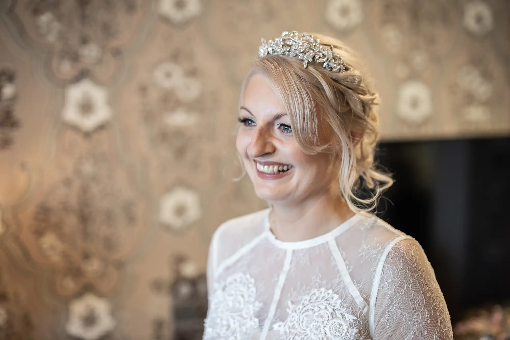 A smiling bride with blonde hair styled in an up-do and decorated with a jewelled tiara, wearing a lace wedding dress with sheer sleeves, stands in front of patterned wallpaper.