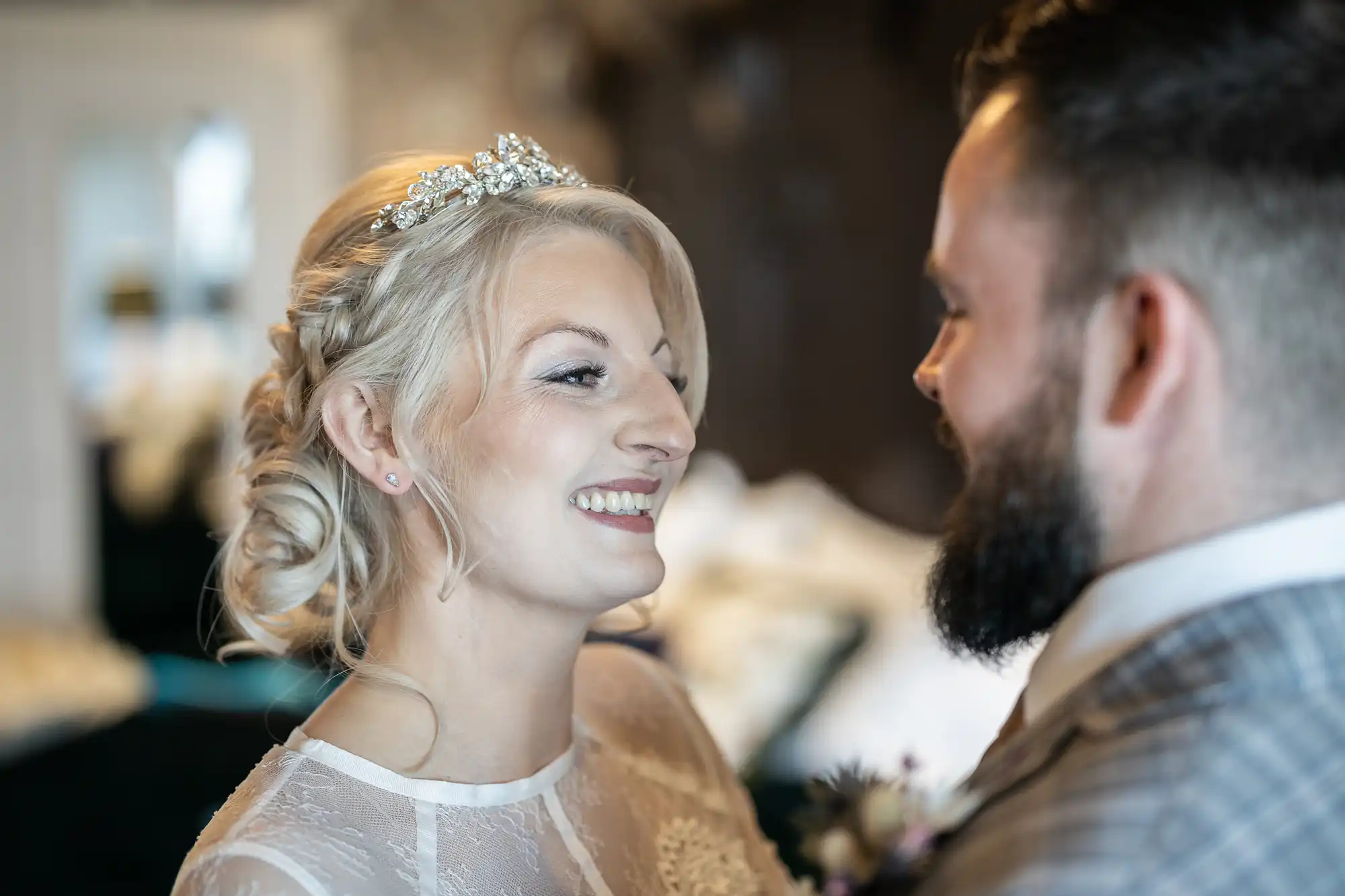 A bride with blonde hair in an updo and jewelled headpiece smiles at a bearded groom in a checked suit. They stand close together indoors, sharing an intimate, joyful moment. The background is softly blurred.