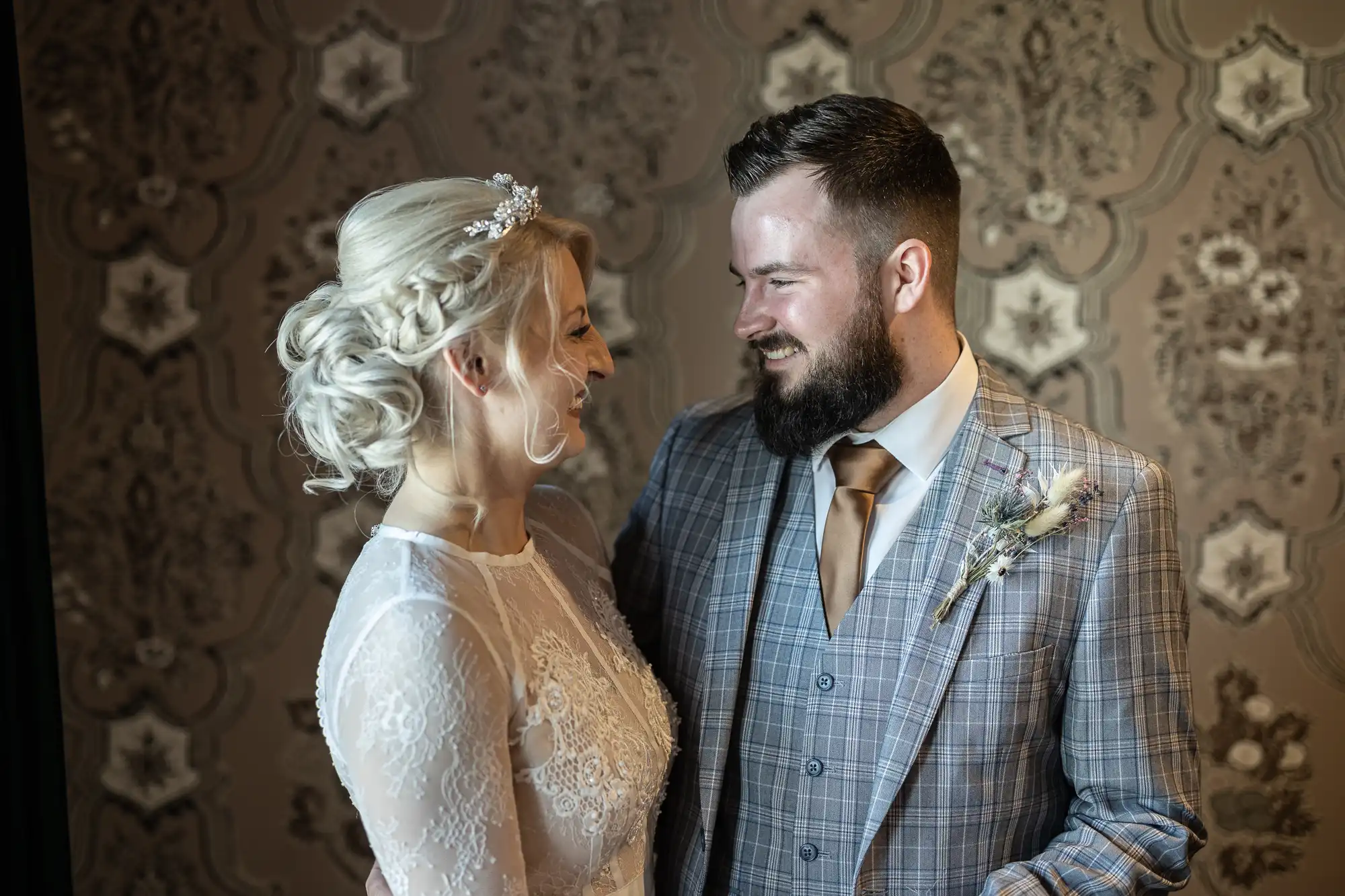 A bride and groom stand close, smiling at each other. The bride has blonde hair styled up and wears a white lace dress and hairpiece. The groom has a beard and wears a grey checked suit with a gold tie. The background is ornate patterned wallpaper.