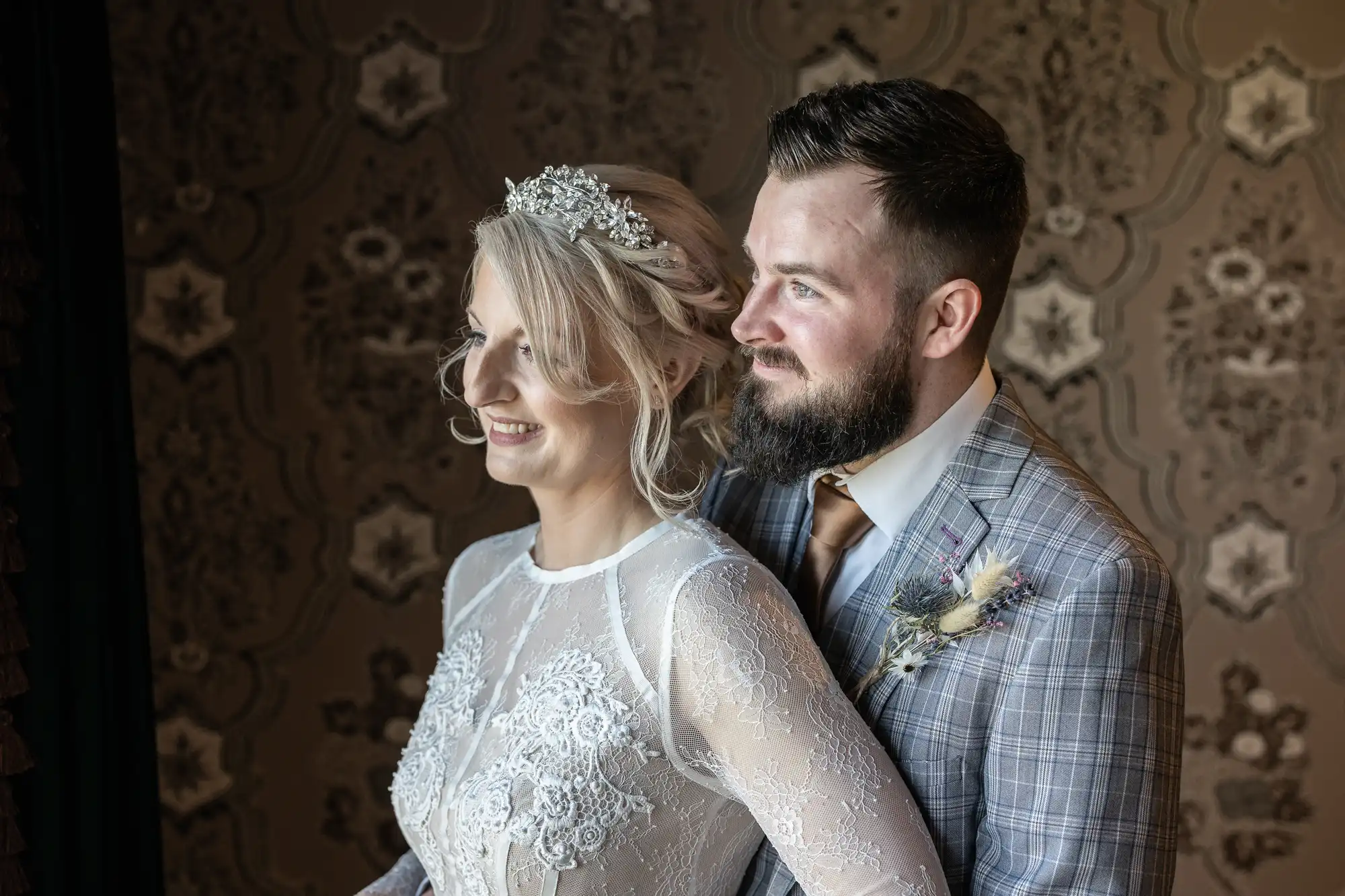 A bride in a white lace wedding dress and jewelled headpiece stands next to a groom in a grey checked suit with a buttonhole. They both smile and gaze to the left, with warm light illuminating them against a vintage, patterned wallpaper background.