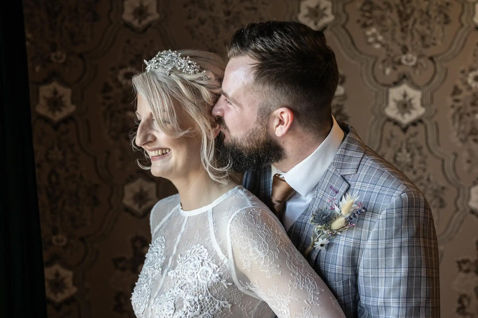 A bride in a lace dress and jewelled headpiece smiles as a groom in a checked suit whispers in her ear. They stand close together against ornate, patterned wallpaper, sharing an intimate, joyful moment.