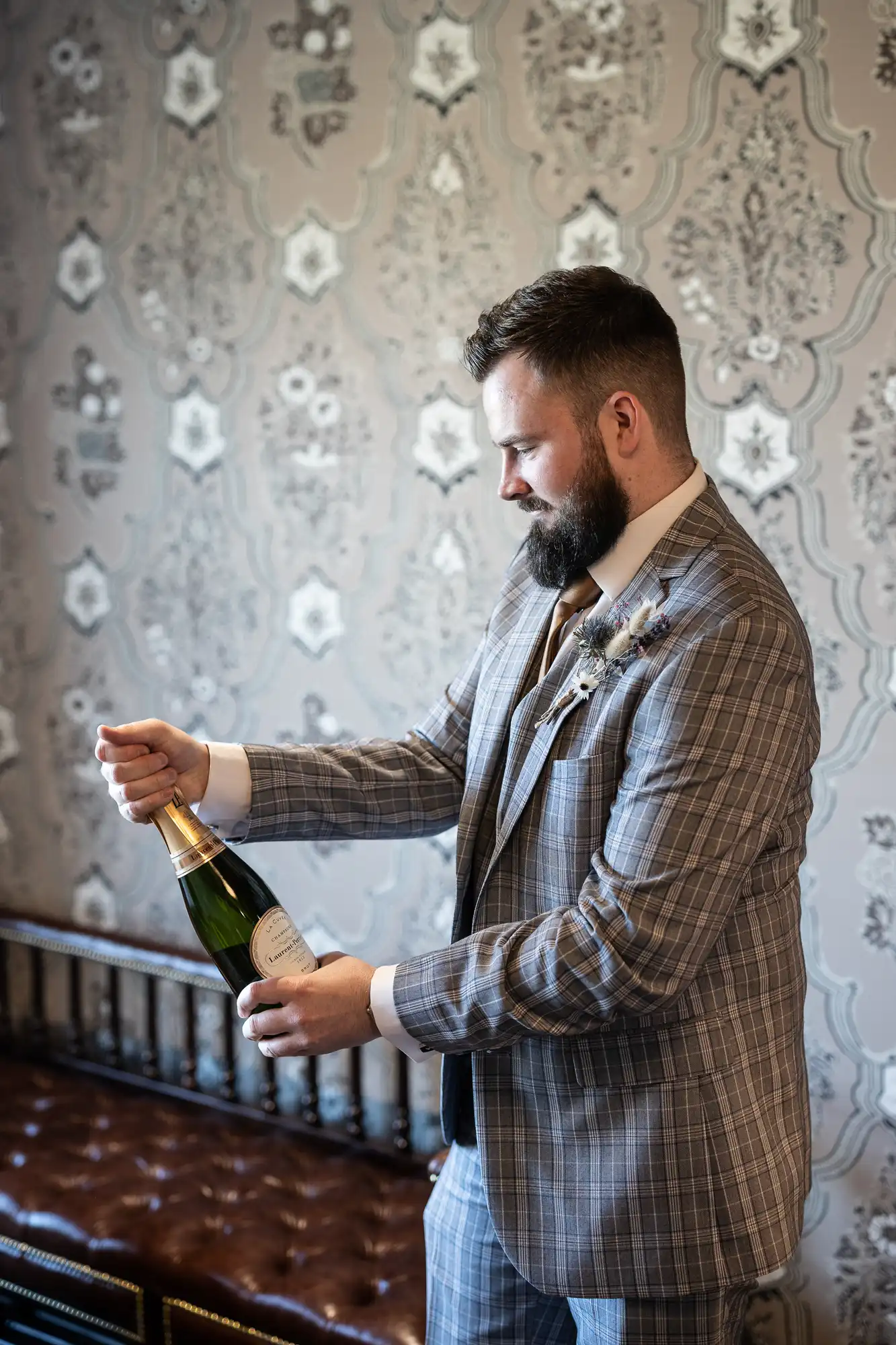 A bearded man in a checked suit stands indoors against ornate wallpaper, smiling as he opens a bottle of sparkling wine. The bottle is held at an angle, and he appears to be preparing to celebrate, with a decorative buttonhole on his lapel.