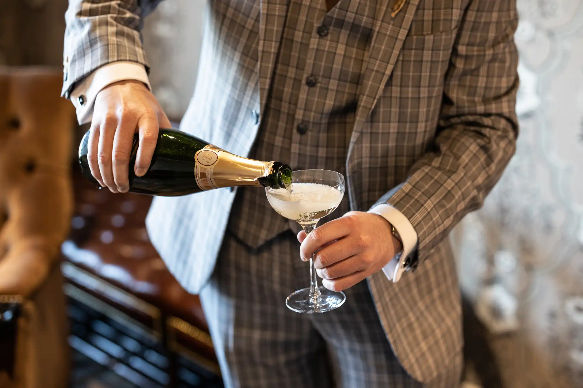A person wearing a grey checked suit pours champagne from a green bottle into a coupe glass. The elegant setting features a buttoned brown chair and patterned wallpaper in the background, suggesting a celebratory or formal occasion.