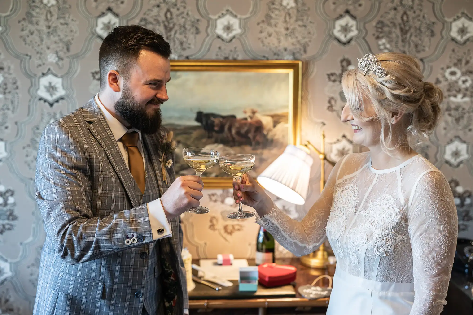 A groom in a checked suit and a bride in a lace wedding dress smile at each other as they clink champagne glasses in a warmly lit room with patterned wallpaper, a lamp, a painting of cows, and a small table with gift boxes and a wine bottle.