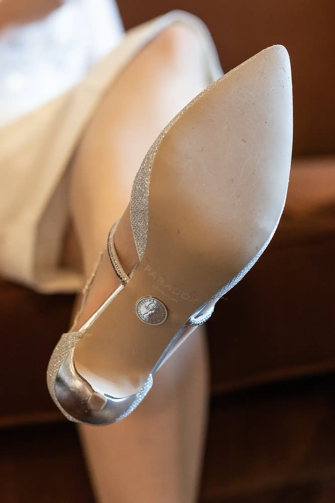 A close-up view of a person’s crossed legs, focusing on the sole of a pointed, silver high-heeled shoe with a strap. The brand “Paradox” and a circular logo are visible on the clean sole. The person is wearing a light-coloured skirt or dress.