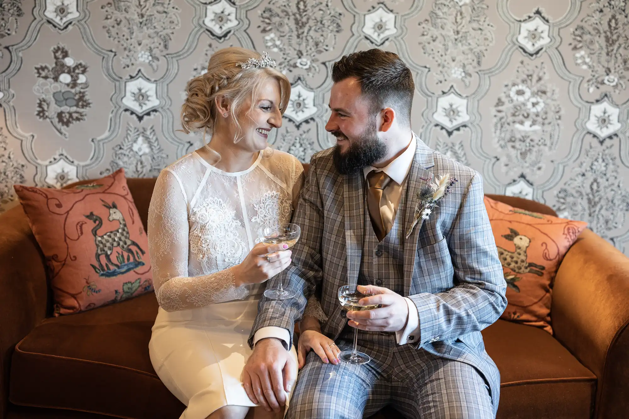 A bride in a white lace dress and a groom in a grey checked suit sit on a brown sofa, smiling at each other and holding champagne glasses. Decorative cushions and ornate wallpaper are in the background, creating an elegant setting.