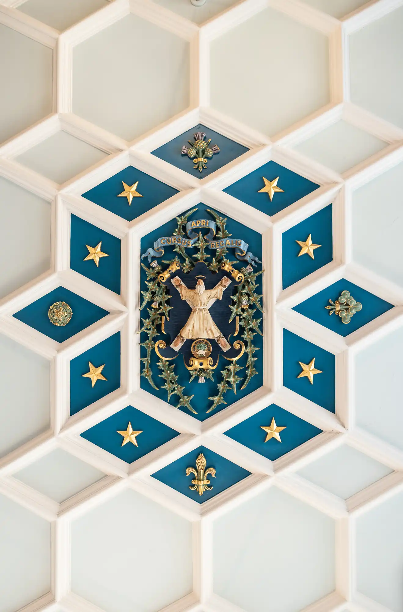 A decorative ceiling with a blue and white hexagonal pattern features gold stars, leaves, and fleur-de-lis. At the centre is a relief of an angel holding a banner inscribed “CARIT HUMILIT SAPIENT,” surrounded by ornate gold embellishments.